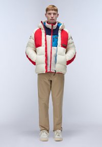 Puffer jacket in cream with red and blue panels, featuring a zipper, hood, and a soft interior. Paired with beige pants and light shoes.