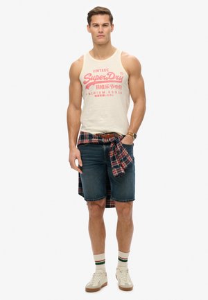 Witte tank top met rode print, blauwe denimshorts, rood-zwart geruit overhemd geknoopt om de taille, witte sneakers met bruine zolen en gestreepte sokken.