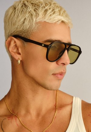 Mladić s kratkom izbjeljenom kosom koji nosi sunčane naočale s crnim okvirom, zlatnu obruč naušnicu, zlatnu lančić ogrlicu i bijelu majicu na bretele.