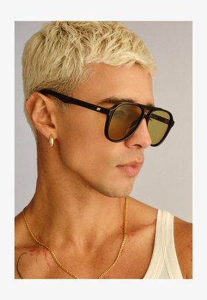 Junge Person mit kurzem gebleichtem Haar, die schwarze Sonnenbrille mit Rahmen, goldenen Creolenohrring, goldene Halskette und weißes Tanktop trägt.