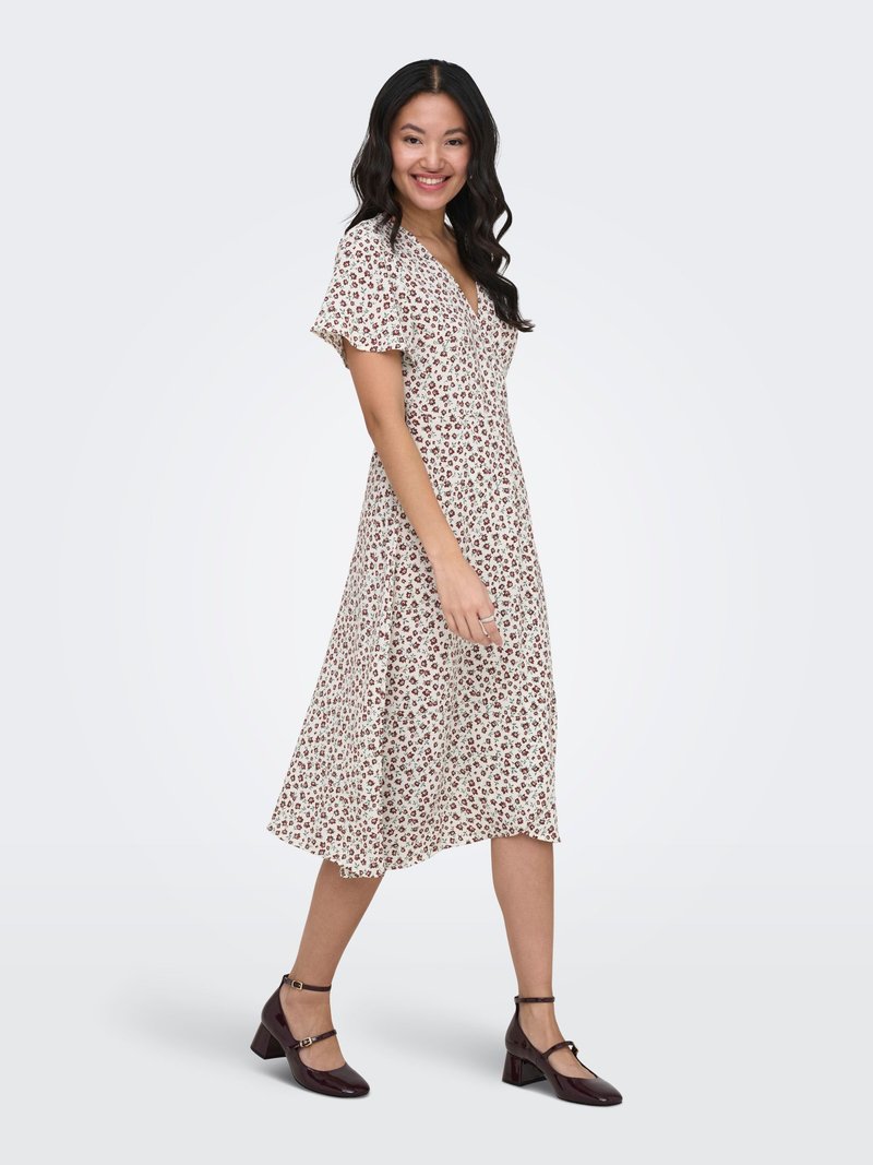 Femme souriante marchant dans une robe midi blanche à imprimé floral rouge avec manches flottantes, portant des talons blocs marron foncé avec bride à la cheville.