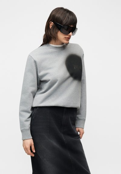 Γυναίκα που φοράει υπερμεγέθη μαύρα γυαλιά ηλίου, γκρι φούτερ με μαύρη λεπτομέρεια κύκλου και μαύρη τζιν φούστα, στέκεται μπροστά σε ένα απλό φωτεινό φόντο.