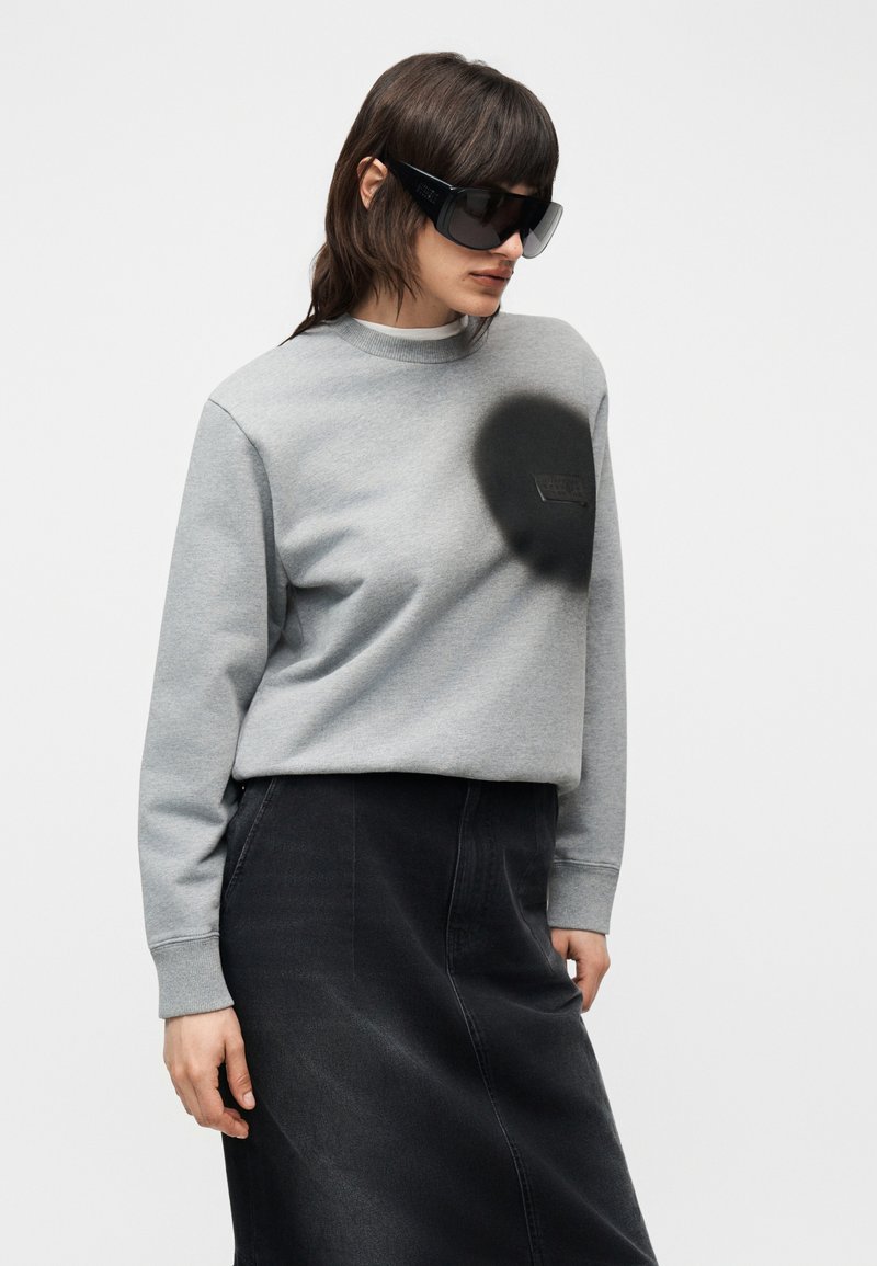 Donna che indossa occhiali da sole neri oversize, felpa grigia con dettaglio a cerchio nero e gonna di jeans nera, in piedi contro uno sfondo chiaro e semplice.