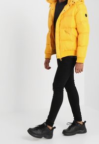 Gelbe Steppjacke mit Kapuze, Reißverschlusstaschen und elastischen Bündchen; kombiniert mit schwarzen Skinny-Jeans und schwarzen Sneakers.