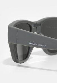 Ochelari de soare gri cu finisaj mat, având lentile întunecate și un logo îndrăzneț "ARMANI EXCHANGE" pe bradă. Design modern, unghiular.
