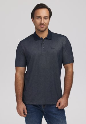 Mężczyzna o krótkich ciemnych włosach, ubrany w ciemnoszarą koszulkę polo z czarnym kołnierzykiem i niebieskie dżinsy, stoi na jasnoszarym tle.