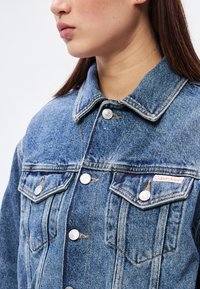 Világoskék farmerdzseki, gallérral, két mellkasi zsebbel fedéllel és ezüst színű fém kiegészítőkkel. A Calvin Klein logó a bal mellrészen található.