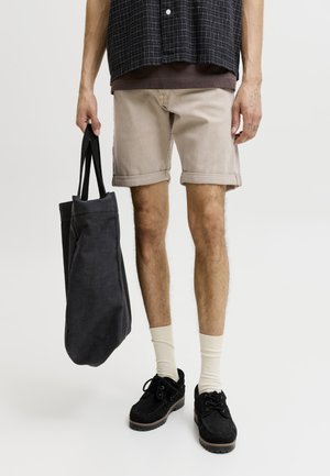 Person i beige shorts, sortternet skjorte, cremefarvede sokker og sorte sko, der holder en stor sort mulepose mod en ensfarvet baggrund.
