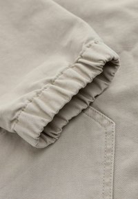 Gros plan sur une manche en tissu beige avec un poignet élastique et des coutures visibles sur un matériau texturé.