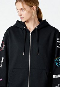 Hanorac negru cu fermoar și șnururi, cu modele grafice albe și albastre pe mâneci, din material moale, și buzunar frontal tip cangur.