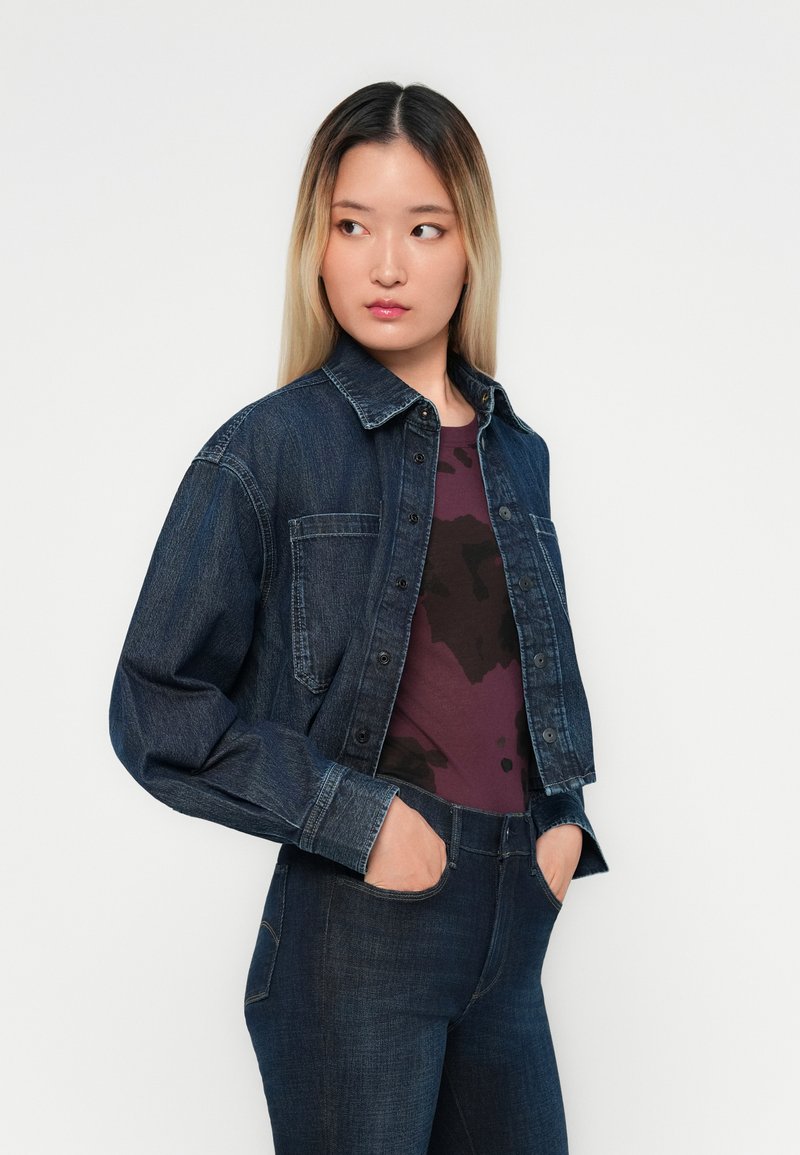 Denimjakke med mørk vask, med knaplukning foran, brystlommer og lange ærmer, båret over en mørkerød top med et mønster.