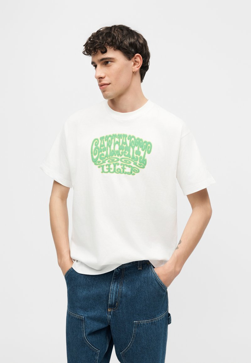 Giovane uomo con capelli ricci scuri, indossa una t-shirt bianca con testo verde stilizzato e jeans blu, mani in tasca, guarda di lato.