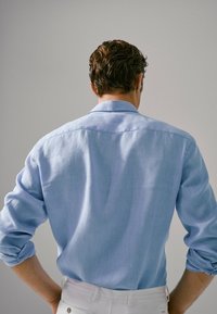 Vaaleansininen pitkähihainen paita, joka on rentoa mallia ja käännetyt hihansuut. Siinä on klassinen kaulus ja hienoinen tekstuuri, esitetty takaa.