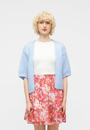 Femme aux cheveux blonds bouclés portant un cardigan ouvert bleu clair, un haut blanc et une jupe rouge à motifs floraux, debout devant un fond clair uni.