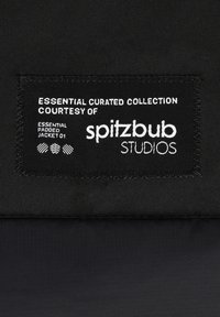 Schwarzes Stoffetikett mit weißem Text: "WESENTLICHE KURATIERTES SORTIMENT COURTESY OF spitzbub STUDIOS WESENTLICHE GEPOLSTERTE JACKE 01."