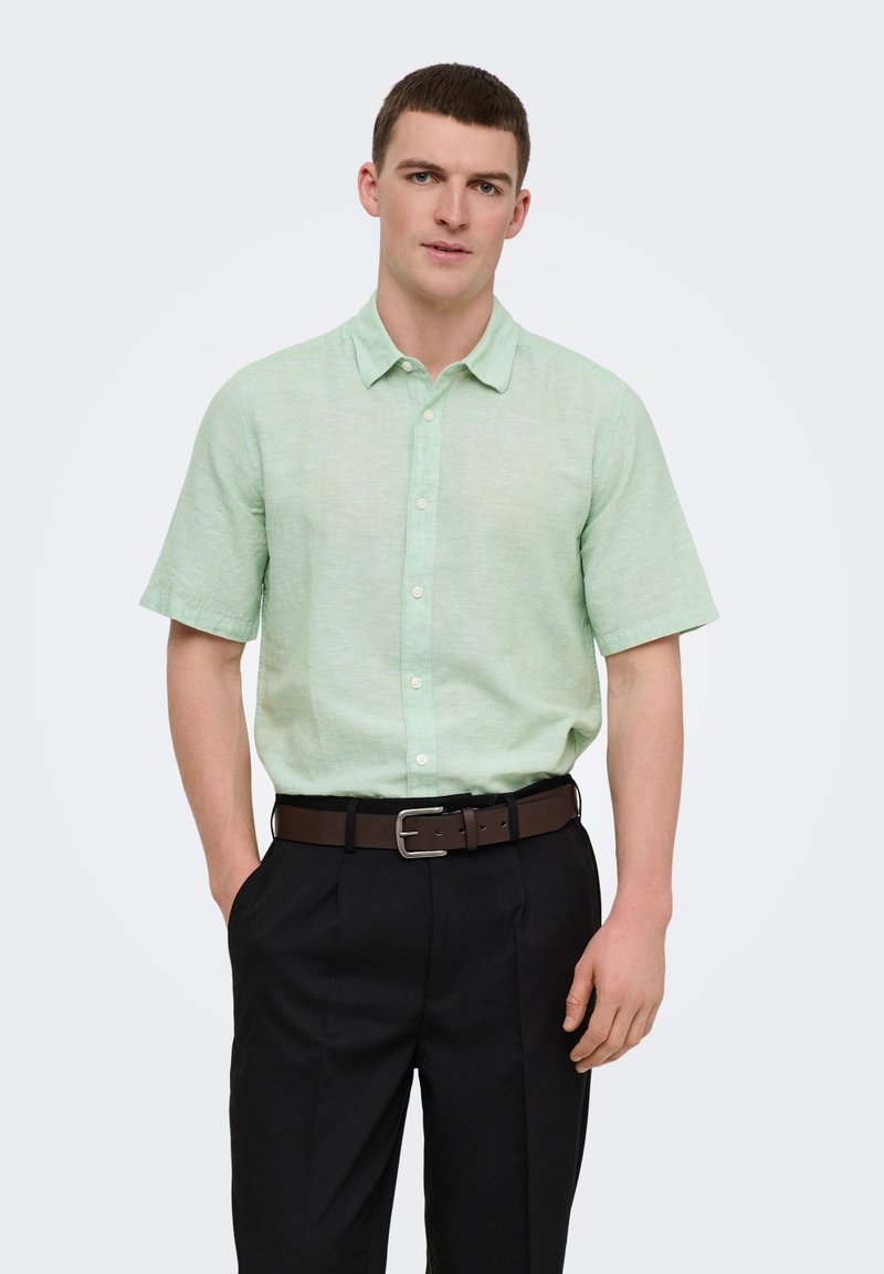 Jeune homme portant une chemise à manches courtes boutonnée vert clair, un pantalon noir et une ceinture marron, debout avec une main dans la poche.