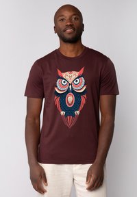 Bordeaux t-shirt met een grote, kleurrijke uilafbeelding met complexe patronen, heldere blauwe en rode accenten. Korte mouwen en een ronde hals.