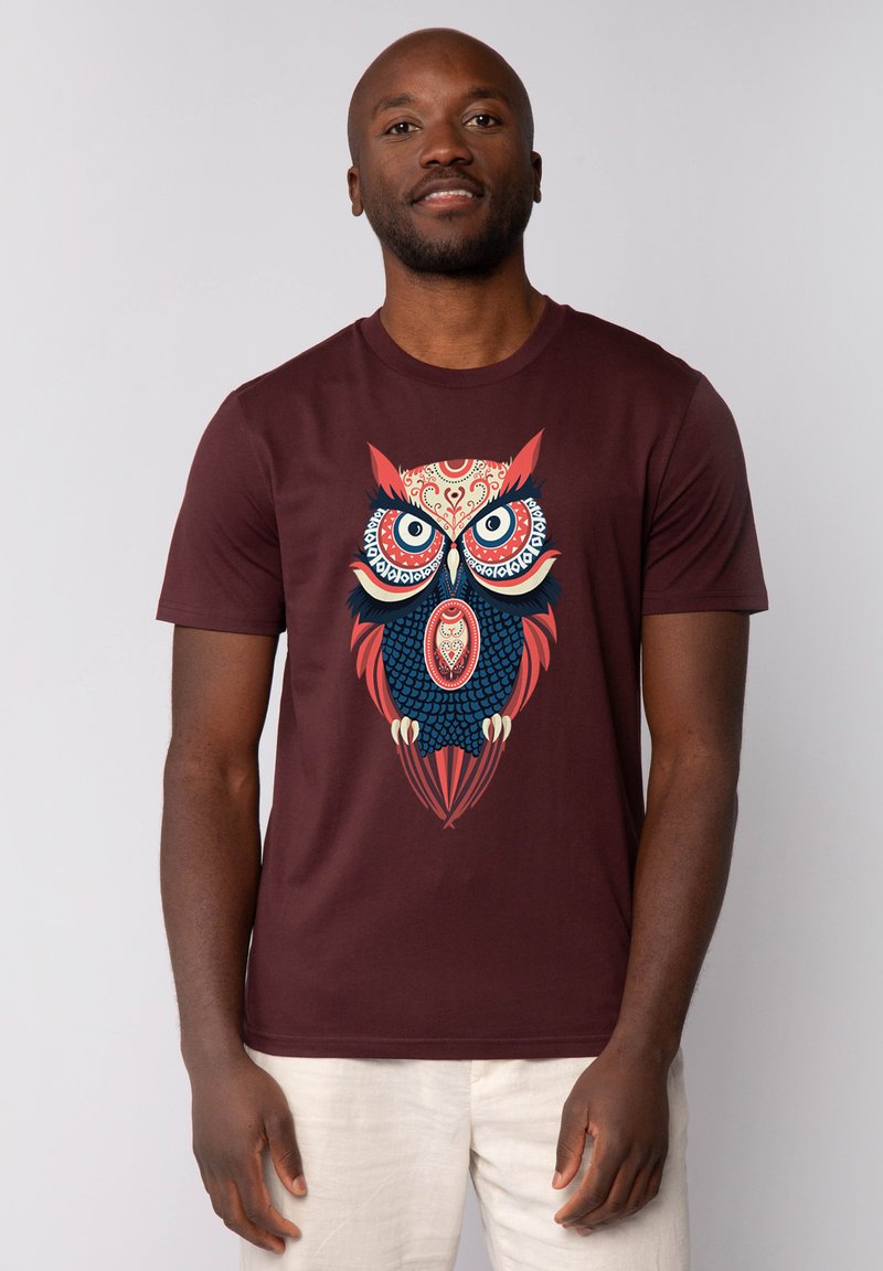 Bordeaux t-shirt met een grote, kleurrijke uilafbeelding met complexe patronen, heldere blauwe en rode accenten. Korte mouwen en een ronde hals.