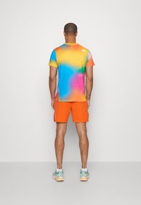 Muž stojící s otočeným zády, oblečený v tričku s multicolor gradientem, jasně oranžových šortkách a modro-bílých teniskách na jednoduchém pozadí.