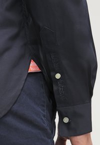 Námořnická košile na knoflíky s hladkou texturou, zdobená kontrastním prošíváním, malým vyšívaným logem na rukávu a značkovou etiketu na lemování.