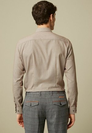 Homme portant une chemise beige à manches longues et un pantalon gris à carreaux avec poches arrière, debout face à un mur beige, dos visible.