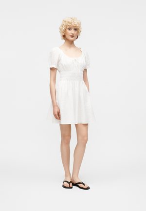 Žena s kučeravými blond vlasmi, oblečená v biele krátke šaty s naberanými rukávmi a čierne žabky, stojí na jednoduchom svetlom pozadí.