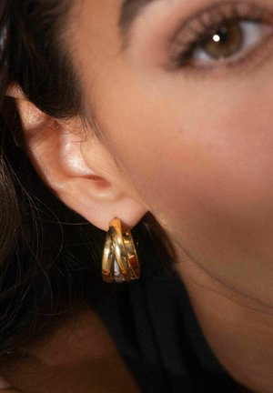Boucles d'oreilles en or et en argent avec un design torsadé, présentant une finition polie et trois bandes entrelacées, accrochées à une oreille.