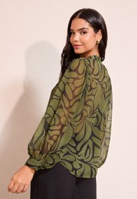 Ženska z dolgimi temnimi lasmi nosi prosojno zeleno bluzo s črnim abstraktnim vzorcem listov in črne hlače, gleda preko ramena in nežno se smehlja.