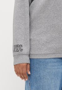 Gray sweatshirt with cuff detail showing "NYC" in black embroidery. Textured fabric with a relaxed fit, paired with blue denim jeans.
