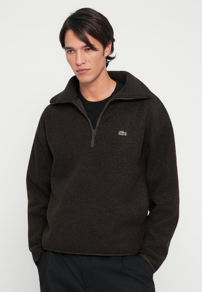 Brun fleece pullover med høj krave, kvart lynlås fortil og lille grønt logo. Tekstureret stof med en afslappet pasform og lange ærmer.