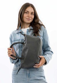 Sac à tote gris avec une texture lisse et des poignées doubles, doté d'une fermeture à glissière et d'un design minimaliste, tenu par une personne portant une veste en denim.