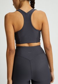Donkergrijze geribbelde sportbeha met brede schouderbanden, voorzien van een racerback-ontwerp. De stof heeft een zachte textuur met subtiele branding op de achterkant.