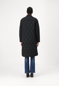 Abrigo largo acolchado negro con corte recto, mangas largas y una abertura trasera, combinado con jeans de mezclilla acampanados y zapatos de plataforma.