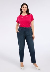 Fuchsia Kurzarmhemd mit floralem Stickmuster und hochgeschnittenen dunkelblauen Jeans. Das Modell trägt spitze weiße Absätze.