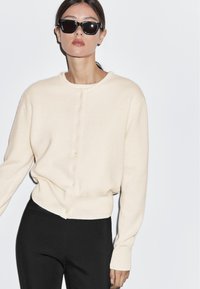 Beige Strickcardigan mit rundem Halsausschnitt, durchgehender Knopfleiste und langen Ärmeln, kombiniert mit schwarzen enganliegenden Hosen.