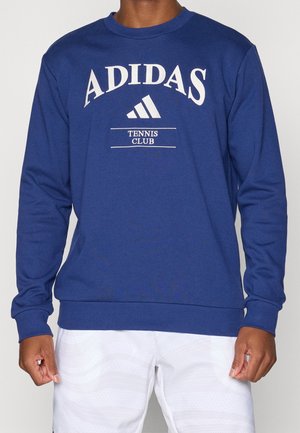 Sweatshirt Adidas bleu à manches longues avec le texte "Tennis Club", porté par une personne visible du cou jusqu'aux hanches.