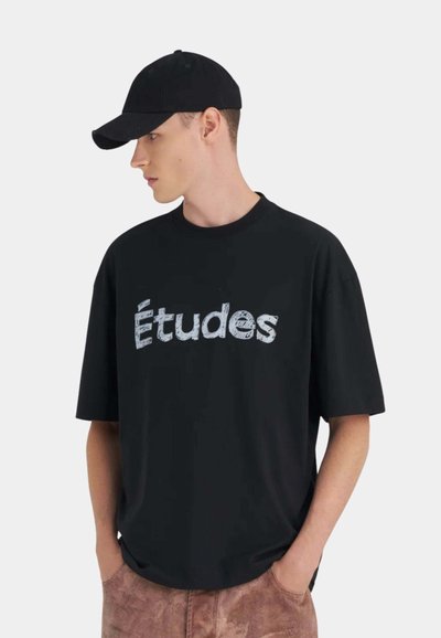 Jeune homme portant une casquette noire et un t-shirt noir oversized avec le texte "Études", debout les mains dans les poches d'un pantalon marron, regardant vers la gauche.