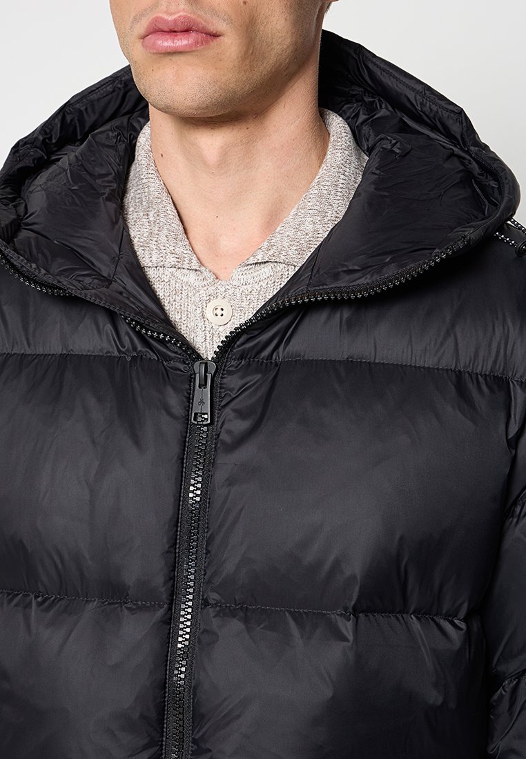 Schwarze Pufferjacke mit Kapuze, glänzender Oberfläche, Reißverschluss vorne und gestepptem Design. Wird über einem hellgrauen Pullover mit Knöpfen getragen.