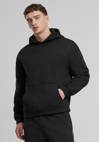 Schwarzer Kapuzenpullover mit einer Fronttasche, aus weichem Stoff gefertigt. Hat einen lockeren Schnitt und gerippte Bündchen. Keine Muster oder Akzente.