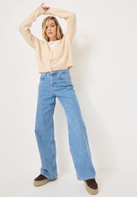 Cardigan en tricot beige avec boutons, associé à un jean large taille haute bleu clair et des baskets à plateforme marron, posé sur fond blanc.