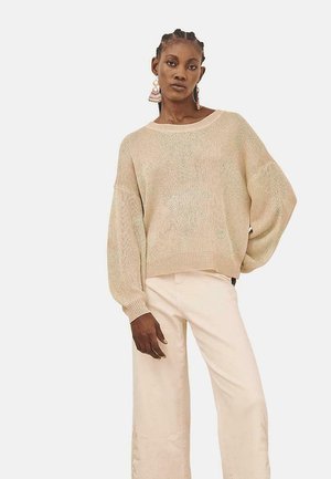 Suéter de punto beige con un ajuste holgado y mangas anchas; presenta sutiles motas verdes, cuello redondo y dobladillo acanalado. Combinado con pantalones claros.