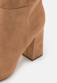 Botín de tobillo de ante marrón con un tacón cuadrado y acabado texturizado, con detalle de costura a lo largo de los bordes.