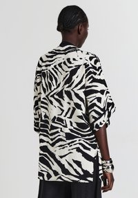 Frau mit geflochtenem Haar trägt ein lockeres schwarz-weißes Zebra gestreiftes Hemd, schwarze Hosen und silberne Armreifen, mit dem Rücken zu einem einfarbigen Hintergrund.