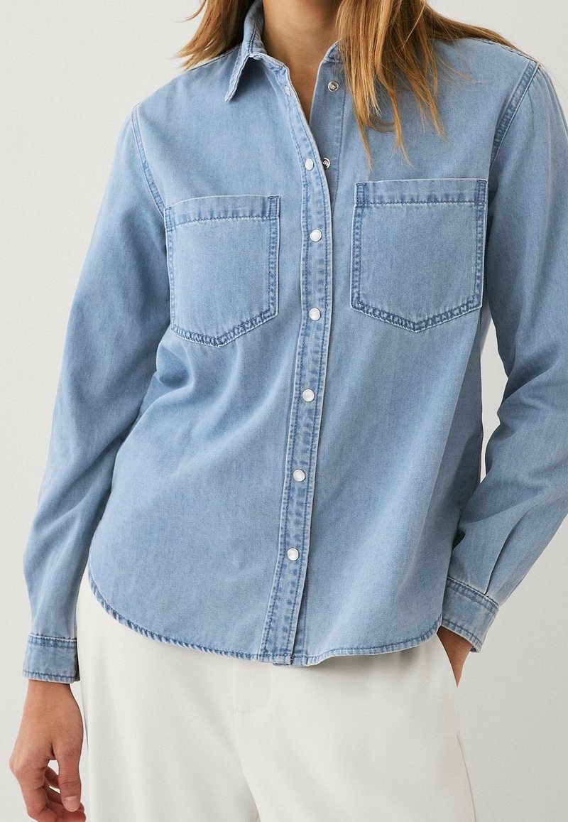 Femme portant une chemise en jean bleu clair boutonnée avec deux poches poitrine et un pantalon blanc sur fond uni.
