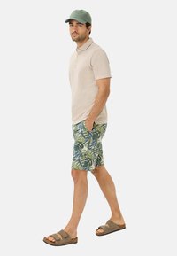 Beige poloshirt, groene pet, gemêleerde groene en blauwe shorts, bruine sandalen, staande zijwaarts met handen in de zakken op een witte achtergrond.