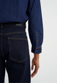 Donkerdenim jeans met oranje stiksels, met een achterzak, leren label, en een close-up van een blauwe lange mouwen shirt met knoopmanschetten.