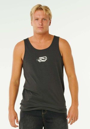 Blonde man met een zwart tanktop met een wit grafisch logo, staand tegen een effen witte achtergrond.