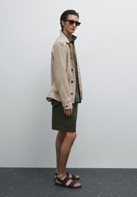 Beige jacket with black buttons, dark green shirt and shorts, brown sandals. Model stands sideways against a neutral background.