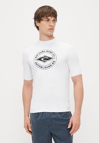 Biała koszulka z krótkim rękawem typu rash guard z czarnym okrągłym logo na piersi. Wykonana z gładkiego, elastycznego materiału. Prosty, sportowy design.