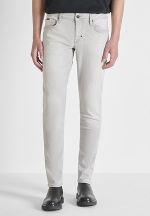 Man draagt lichtgrijze slim-fit jeans en zwarte leren enkellaarzen, staand tegen een effen witte achtergrond.